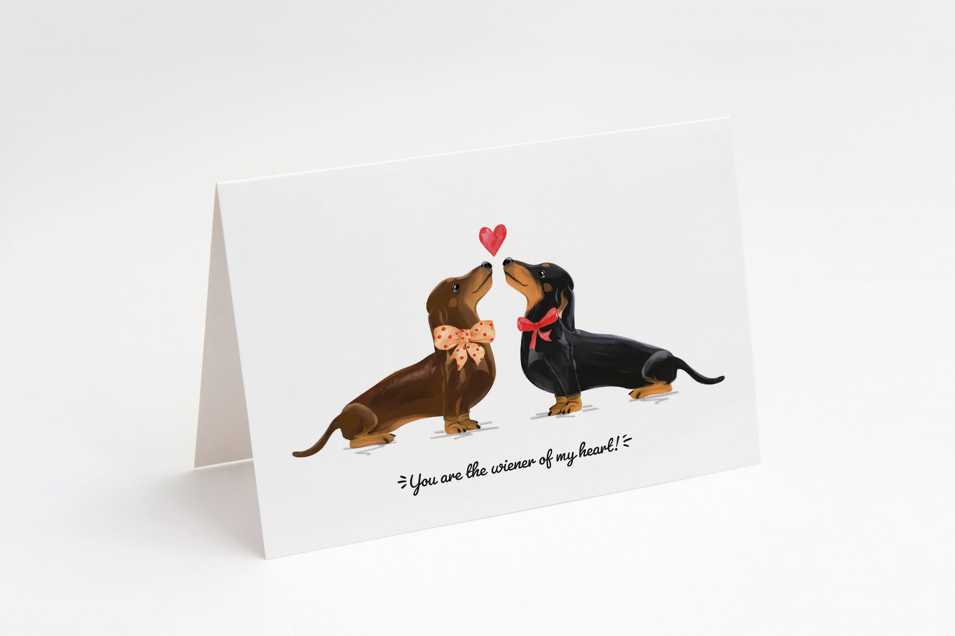 Greeting card with a brown and a black dachshund and a heart in the middle on a white background. "You are the wiener of my heart!" text at the bottom.