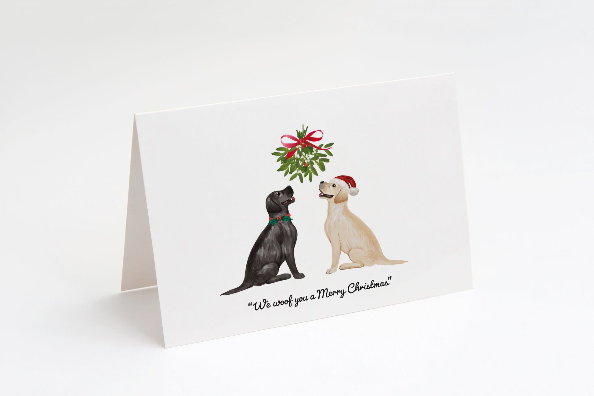 Christmas card with a black and a cream Labrador dog sitting underneath a mistletoe wreath on a white background, with the text: "We woof you a Merry Christmas"