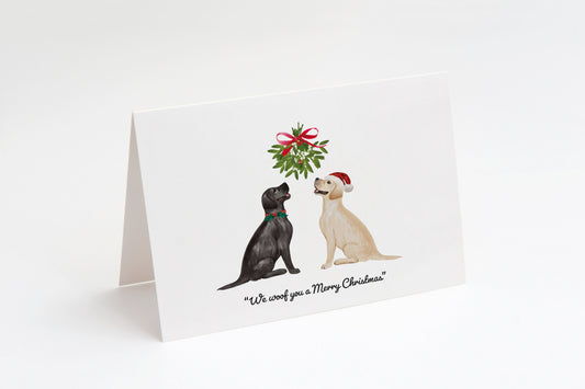 Christmas card with a black and a cream Labrador dog sitting underneath a mistletoe wreath on a white background, with the text: "We woof you a Merry Christmas"