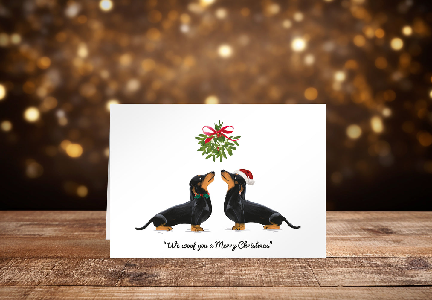Christmas card with two black Dachshund dogs underneath mistletoe on a white background with the text "We woof you a Merry Christmas" underneath. 