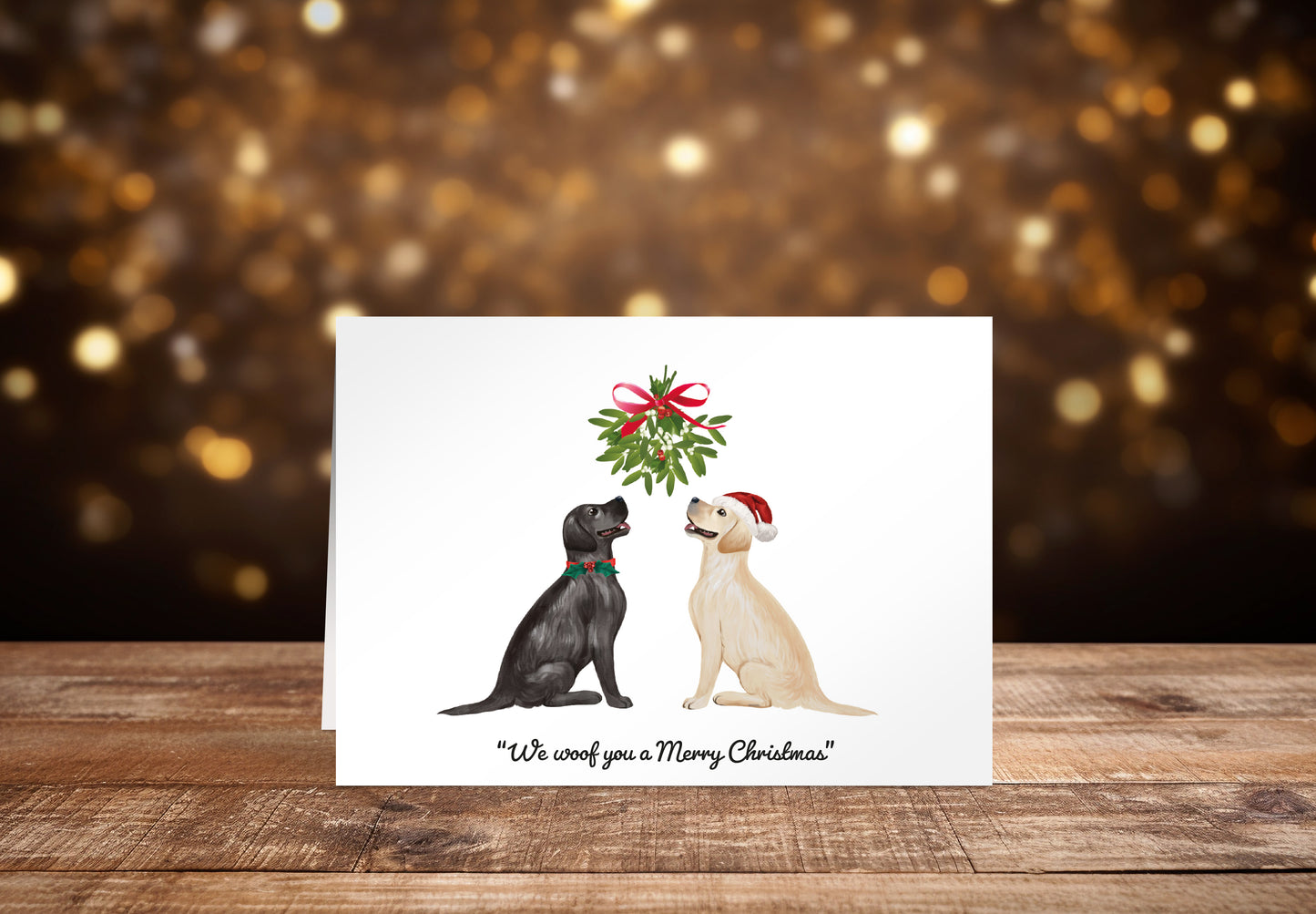 Christmas card with a black and a cream Labrador dog sitting underneath a mistletoe wreath on a white background, with the text: "We woof you a Merry Christmas"