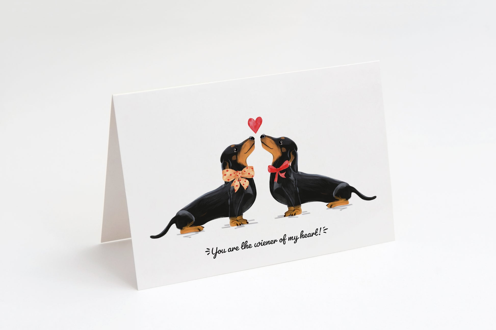 Greeting card with two black dachshunds and a heart in the middle on a white background. "You are the wiener of my heart!" text at the bottom.