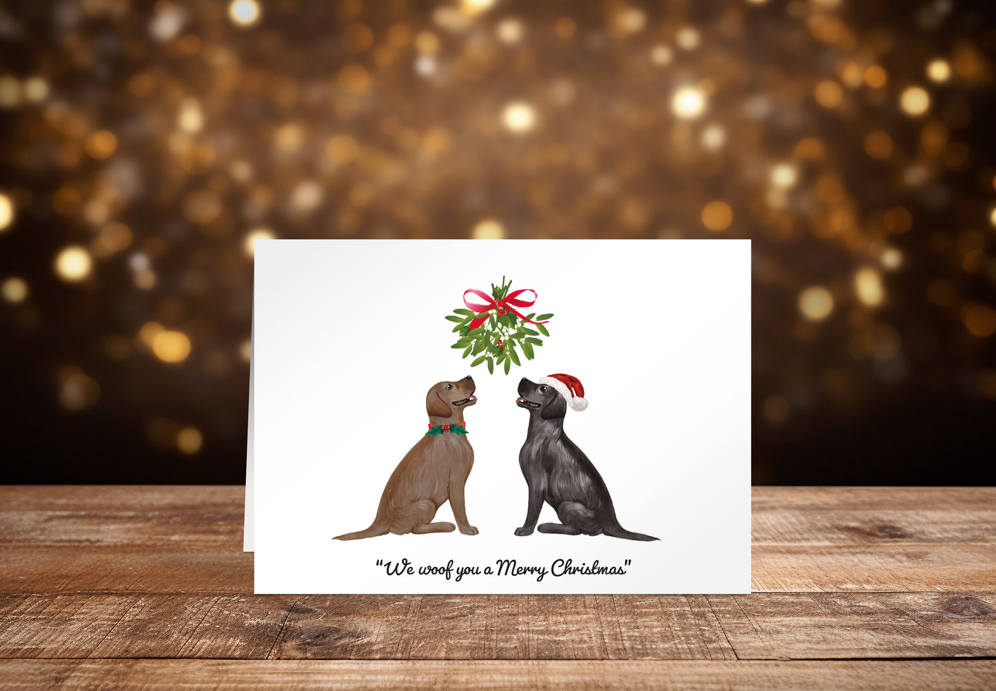 Christmas card with a black and a brown Labrador dog sitting underneath a mistletoe wreath on a white background, with the text: "We woof you a Merry Christmas"