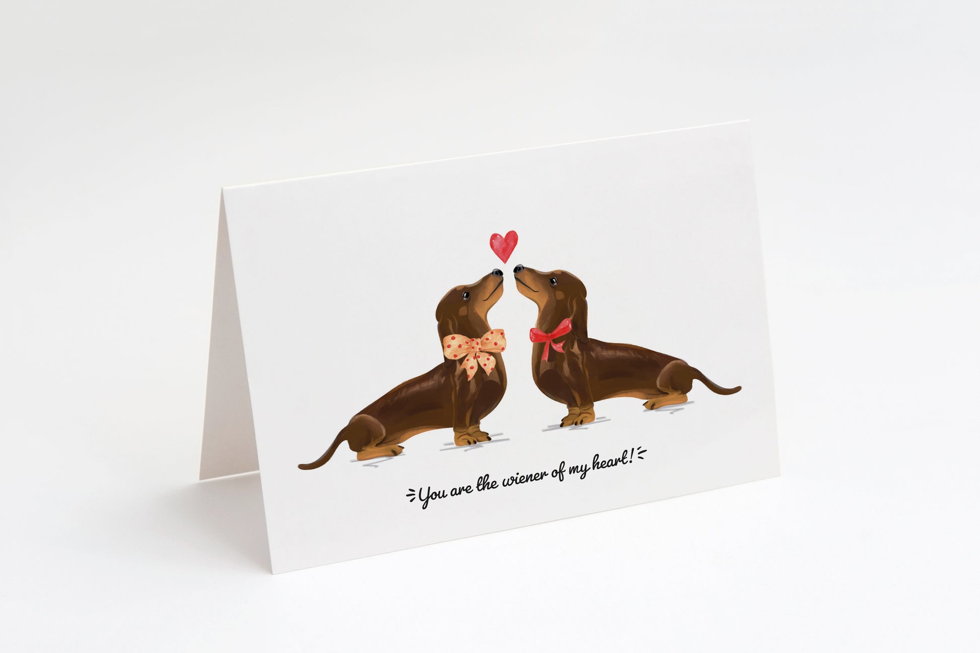 Greeting card with two brown dachshunds and a heart in the middle on a white background. "You are the wiener of my heart!" text at the bottom.
