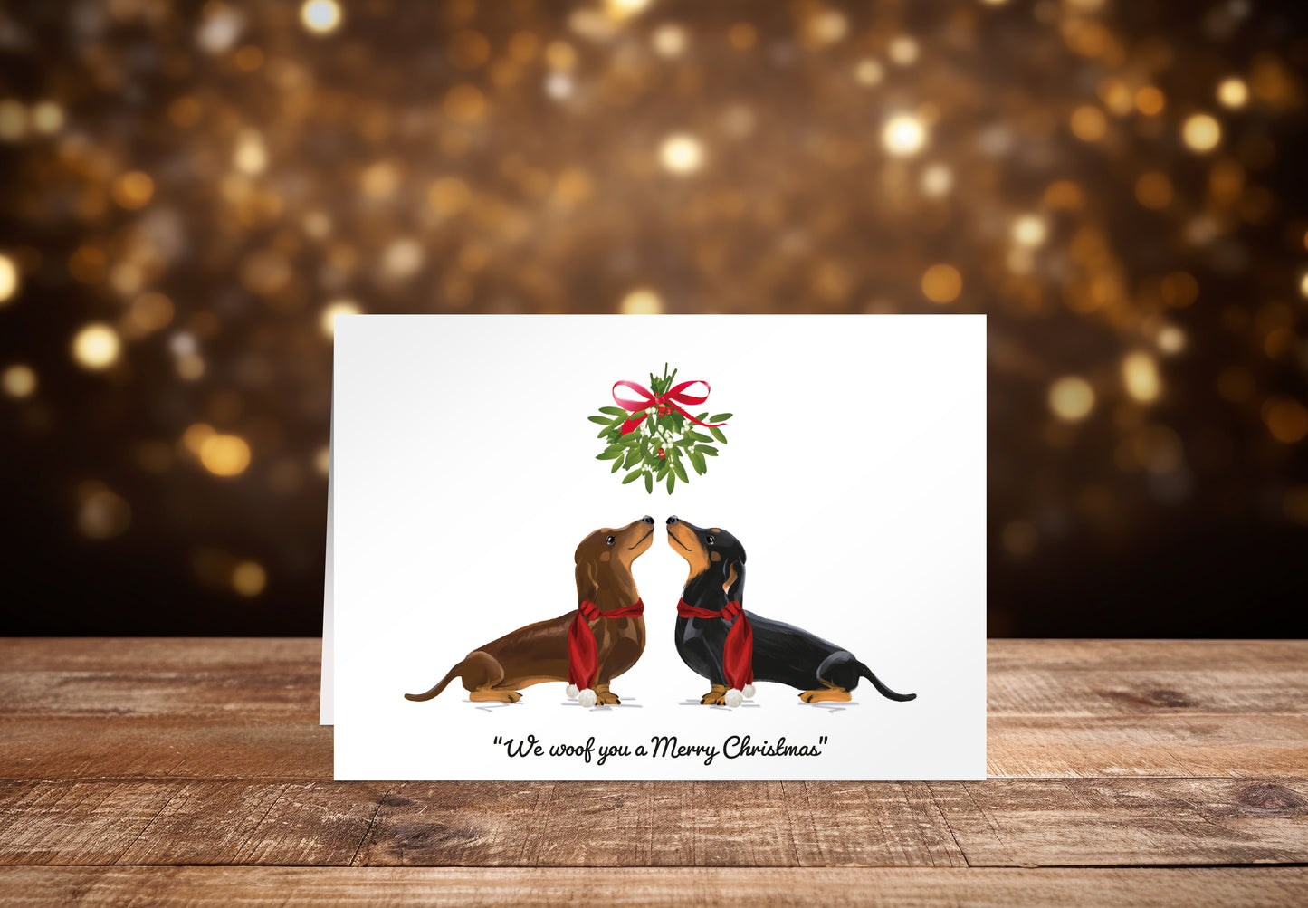 Christmas card with a brown and a black Dachshund dog underneath mistletoe on a white background with the text "We woof you a Merry Christmas" underneath. 