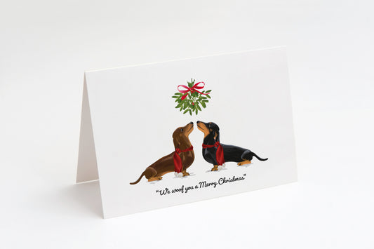 Christmas card with two Dachshund dogs underneath mistletoe on a white background with the text "We woof you a Merry Christmas" underneath. 