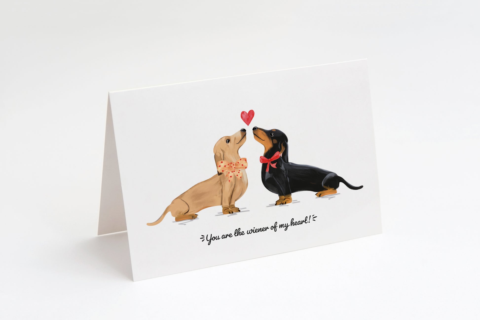 Greeting card with a cream and a black dachshund and a heart in the middle on a white background. "You are the wiener of my heart!" text at the bottom.