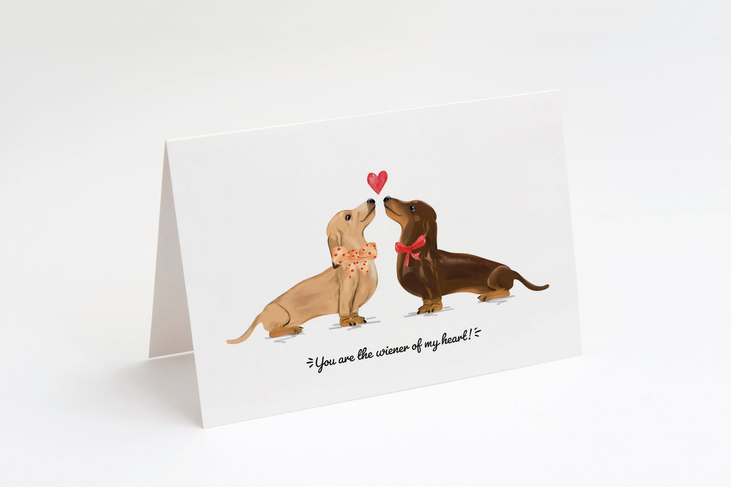 Greeting card with a cream and a brown dachshund and a heart in the middle on a white background. "You are the wiener of my heart!" text at the bottom.