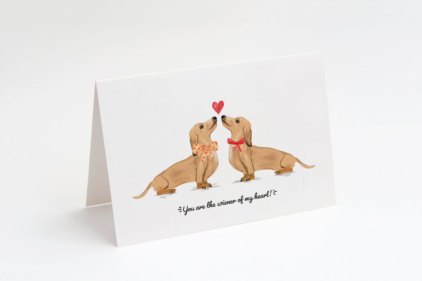 Greeting card with two cream dachshunds and a heart in the middle on a white background. "You are the wiener of my heart!" text at the bottom.