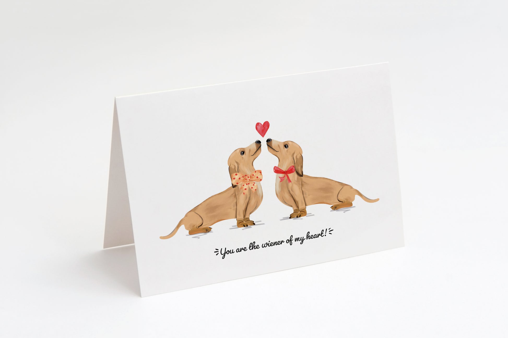 Greeting card with two cream dachshunds and a heart in the middle on a white background. "You are the wiener of my heart!" text at the bottom.