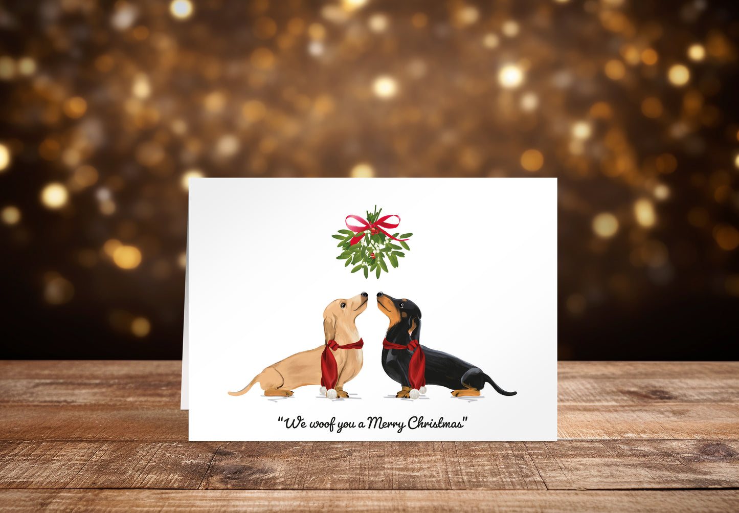 Christmas card with a cream and a black Dachshund dog underneath mistletoe on a white background with the text "We woof you a Merry Christmas" underneath. 