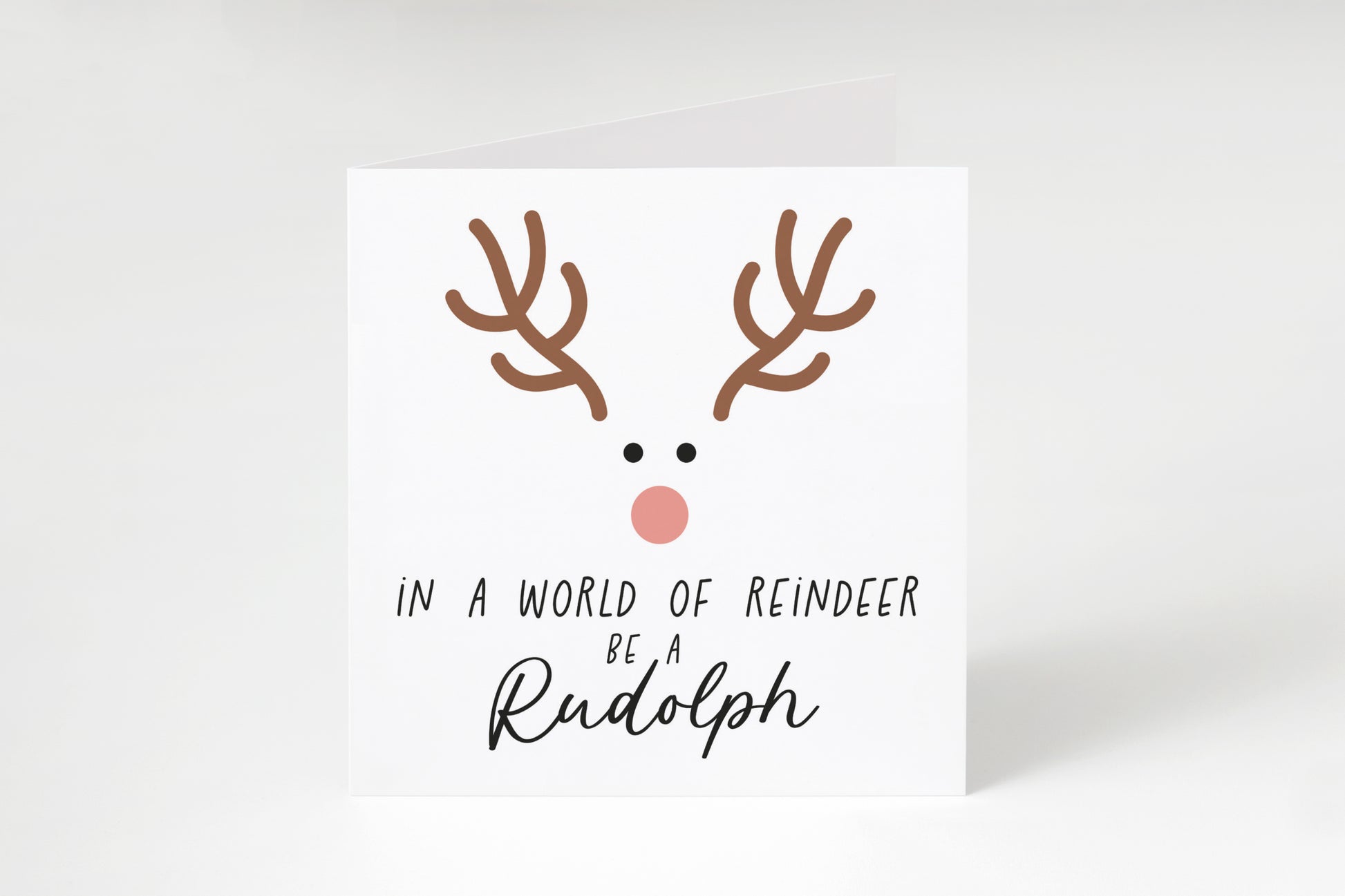 Reindeer face with the text "In a world of reindeer be a Rudolph" on a white square greeting card.
