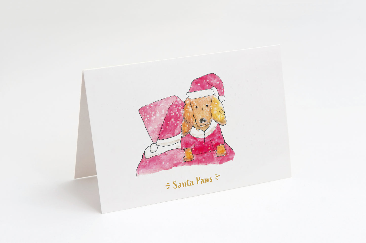 Greeting card with a dog wearing a Santa outfit and hat, sat on Santa's shoulders. Santa is facing away from us. Snow in foreground on a white background.