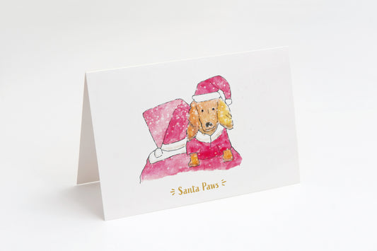 Greeting card with a dog wearing a Santa outfit and hat, sat on Santa's shoulders. Santa is facing away from us. Snow in foreground on a white background.